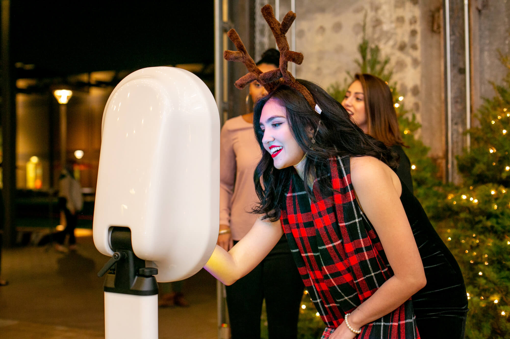 Woman in plaid scarf wearing reindeer antlers using Ozark Pix photo booth to take picture in front of holiday trees